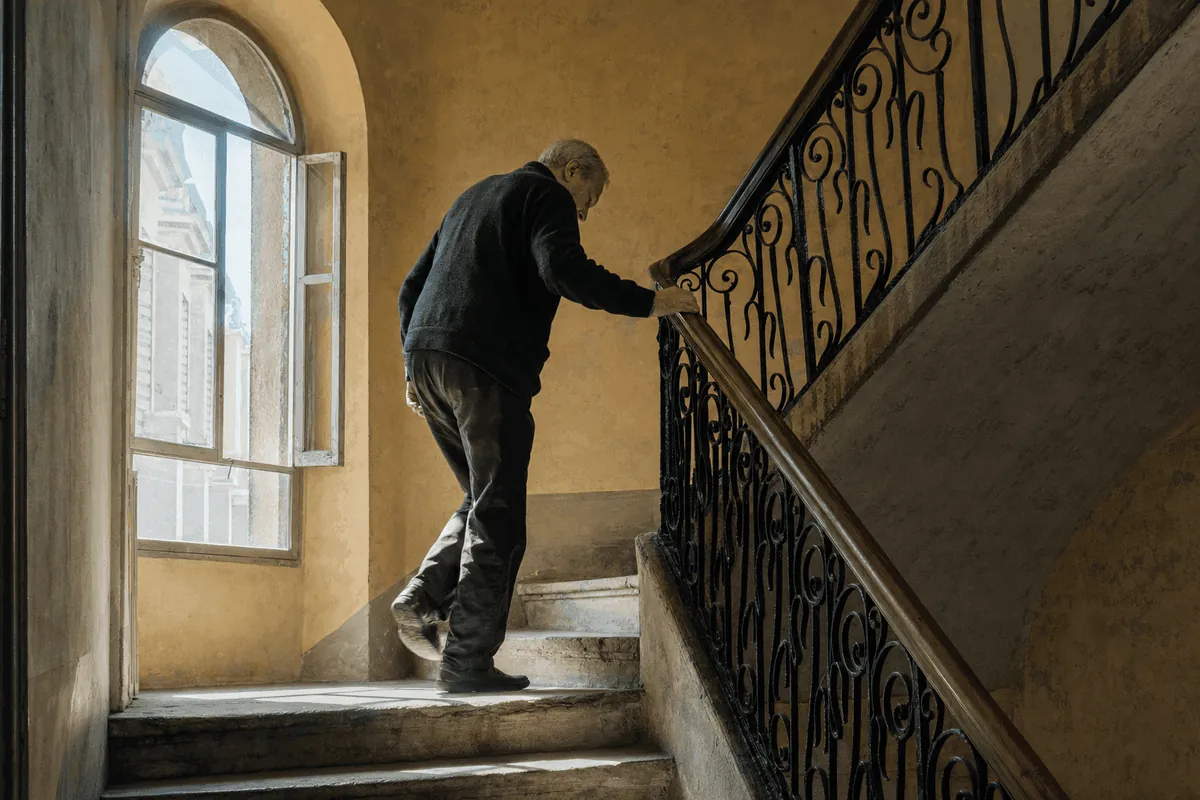 Homme d'une soixantaine d'années sur le palier d'une cage d'escalier en pierre, la main sur la rampe en fer, lumière du matin